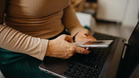 Zwei Hände halten ein Smartphone über der Tastatur eines aufgeklappten Laptops, die Person sitzt in einem beigen Pullover auf dem Sofa
